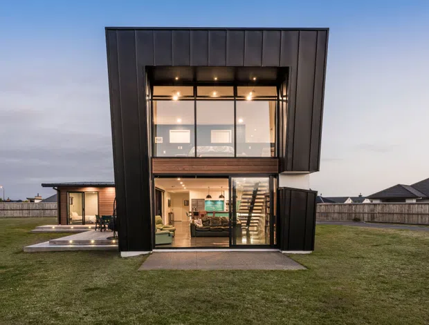 Front exterior view of a contemporary two-storey residence at dusk, featuring a tall projecting central volume clad in dark vertical panels, extensive black-framed glazing, and a glazed ground-floor living area. A smaller single-storey wing and paved terrace extend to the side, with a broad lawn and suburban homes in the background.