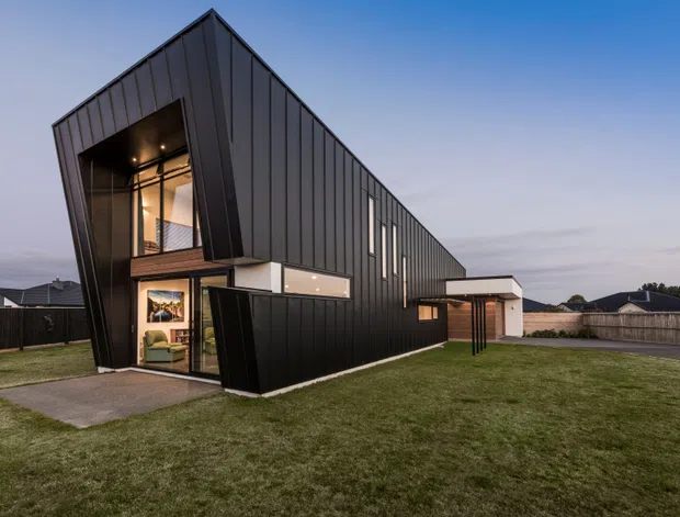 Exterior dusk view of a distinctive contemporary residence with a long, narrow gabled form, dark vertically seamed metal cladding, extensive glazing, and a recessed double-height entry. The home sits on a broad lawn in a suburban setting, with warm interior lighting visible through the entry glazing.