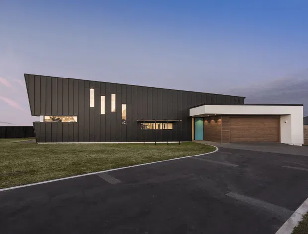A dusk exterior view of a contemporary single-storey residence with a dramatic dark vertical-clad facade, asymmetrical sloping roofline, tall narrow windows, and an attached double garage. A curved driveway leads across the foreground, with lawn surrounding the house.