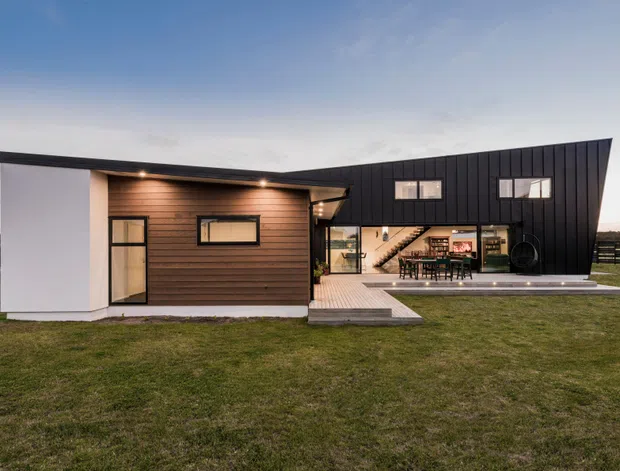 Exterior view of a contemporary two-storey residence with a dramatic asymmetrical sloping roofline and dark vertical cladding. The lower volume combines white rendered walls with warm horizontal timber cladding, while a timber deck leads to a glazed dining and living area visible through the open facade. Set in a broad grassy suburban or rural landscape at dusk.