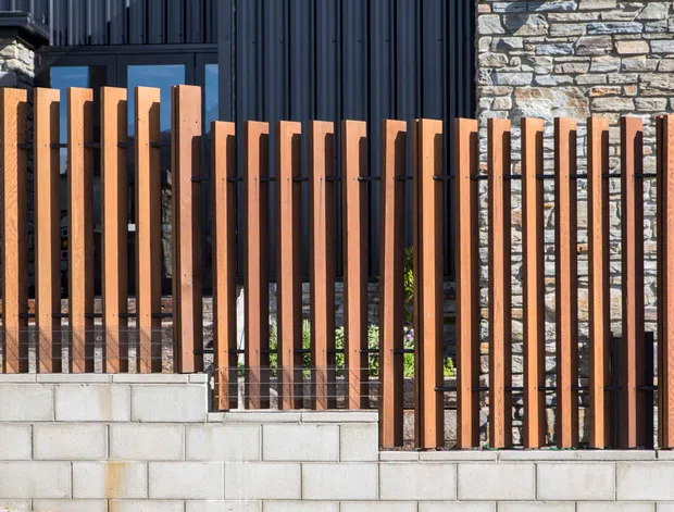 Close-up exterior view of a contemporary residential facade and boundary fence. Vertical warm timber slats are spaced across a dark metal frame, with fine wire infill and a low light-grey concrete block retaining wall. Behind the fence, dark vertical metal cladding, glazing, and rough stacked-stone walling create a layered modern facade.