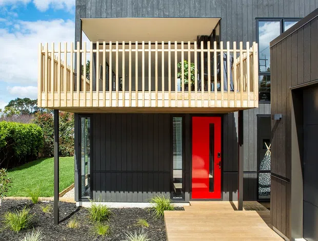Exterior view of a contemporary two-storey dwelling with dark vertical cladding, a projecting upper-level balcony enclosed by pale timber vertical battens, large glazed openings, and a vivid red entry door. A paved walkway, landscaped garden, and attached dark garage volume are visible.