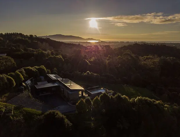 Aerial view of a contemporary residence nestled into a heavily wooded, elevated landscape. The low-profile home has flat roof planes, dark exterior surfaces, and extensive glazing or open terraces, with a broad sunset-lit horizon and distant water visible beyond the surrounding hills.