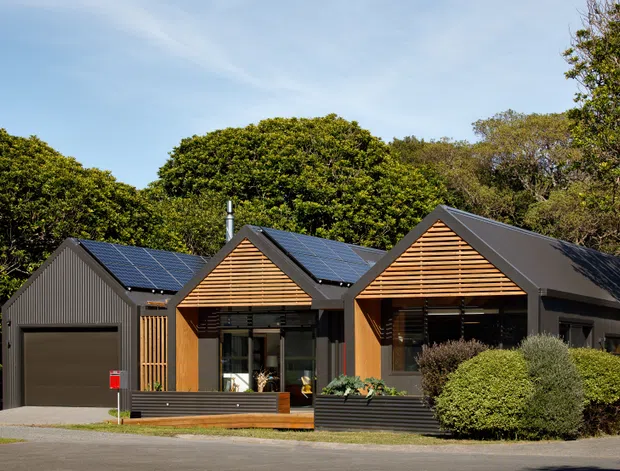 Exterior view of a contemporary single-storey residential building composed of several gabled forms. The dark metal-clad facade is paired with warm horizontal timber screening and large glazed openings, with solar panels visible on the pitched roofs. A garage, entry deck, low planters, and established landscaping sit along the street frontage.