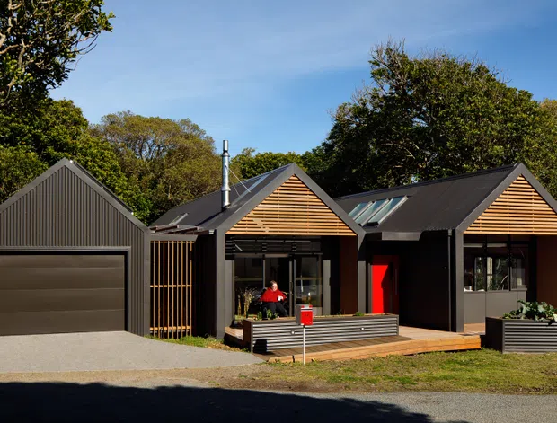 Exterior view of a compact contemporary house composed of several connected gabled volumes. The dark grey vertically clad facade and pitched roofs contrast with warm horizontal timber slat gable screens, large glazed openings, a bright red entry door, and a timber deck. A matching enclosed garage sits to the left, with mature trees surrounding the property.