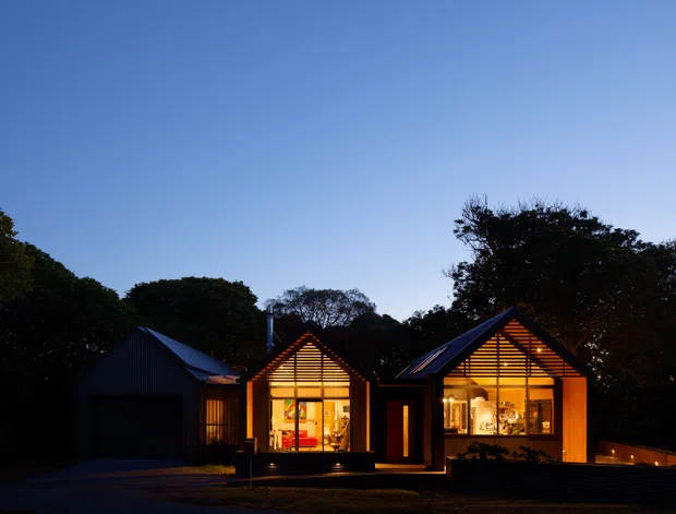 Twilight exterior view of a contemporary single-storey residence composed of several compact gabled pavilions. The dark-clad facade and sharply pitched roofs frame warmly illuminated glazed living spaces, with a recessed entry and attached garage visible in the shadowed left wing. Dense mature vegetation surrounds the property, and low boundary walls and paving define the foreground.