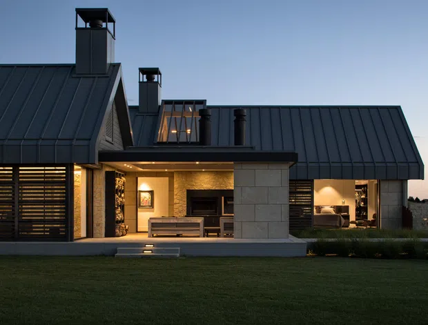 Exterior view of a contemporary single-storey residence at dusk, featuring dark standing-seam pitched roofs, multiple chimney structures, charcoal horizontal screening, warm-lit floor-to-ceiling openings, and a sheltered central outdoor living area with a fireplace. The home sits behind a broad lawn with landscaped planting.