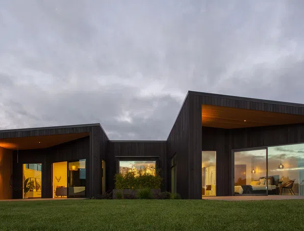 Single-storey contemporary house exterior photographed from the front, with dark vertical timber cladding, strong projecting box-like volumes, recessed entry and large glazed living spaces glowing with warm interior lighting. A broad lawn and overcast sky frame the minimalist facade.
