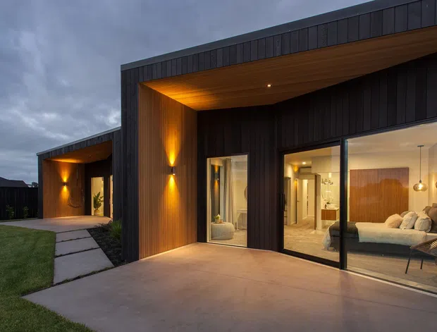 Contemporary single-storey house exterior at dusk, featuring dark vertical cladding, broad flat roof overhangs, warm timber soffits, recessed and wall-mounted lighting, and expansive black-framed glazing. A concrete terrace and stepping-stone path lead across a lawn, with a bedroom visibly furnished behind the main window.
