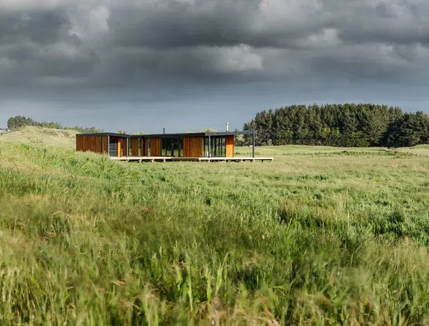 A low, single-storey modern pavilion set within a broad grassy rural landscape. The building has extensive glazing, dark timber or brown cladding, slender structural framing, and a shallow flat roof. Dramatic overcast skies and a dense tree line form the background.