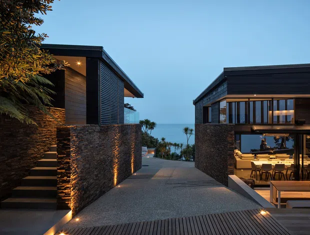 Twilight exterior view of a contemporary coastal residence arranged as two dark-clad pavilion-like volumes around a central pedestrian approach. Rough stacked-stone retaining walls flank the path, with integrated step lighting, timber decking, louvred screening, extensive glazing, and a glimpse of the sea beyond. The right-hand building reveals a warmly lit open-plan dining and living area through large windows.