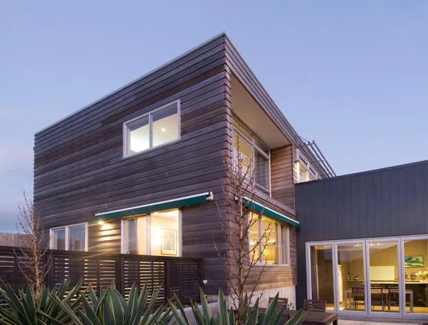 A contemporary two-storey residential exterior at dusk, featuring dark horizontal timber cladding, a projecting upper volume, large windows, slim metal balcony or roofline elements, and an adjoining dark-clad lower structure with glazed doors. Landscaping and spiky plants occupy the foreground.