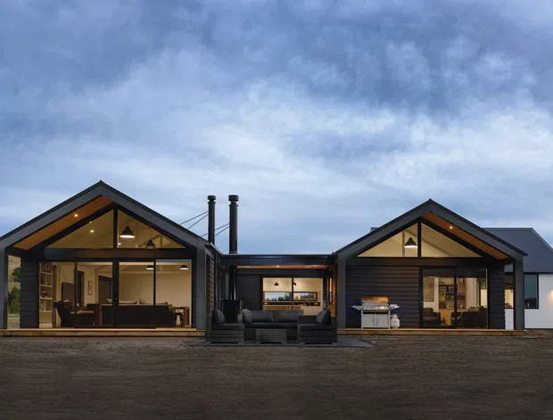 Front exterior view of a low-slung contemporary rural home composed of two dark-clad gabled pavilions linked by a central section. Large glazed openings reveal warm timber interiors, while exposed timber gable framing, black metal cladding, roof forms, and twin chimneys define the facade. The house sits on an open rural site under an overcast sky.