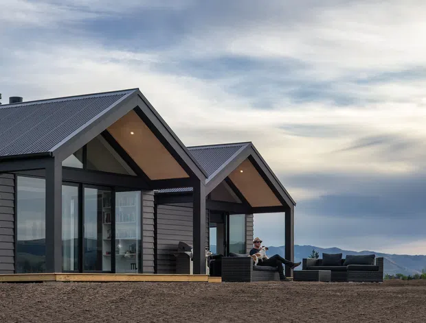 Exterior view of a contemporary multi-gabled home with dark horizontal cladding, charcoal metal pitched roofs, and large glazed openings. Warm timber-lined gable soffits and an outdoor seating area face a broad mountainous landscape.