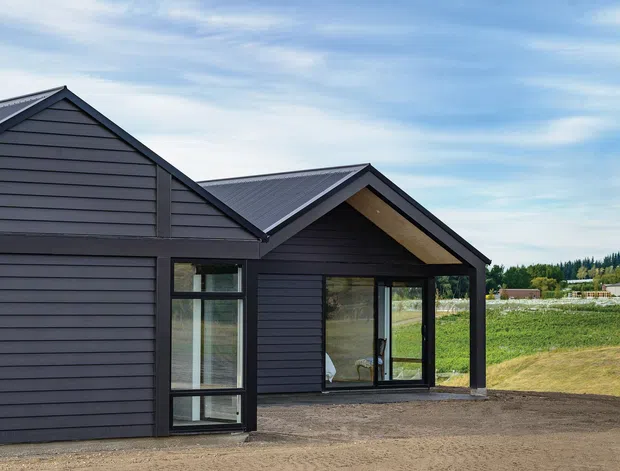 Exterior view of a modern single-storey gable-roof house with dark horizontal cladding, charcoal metal roofing, large glazed openings, and a timber-lined gable soffit. The building sits in a semi-rural landscape with open fields and distant trees.