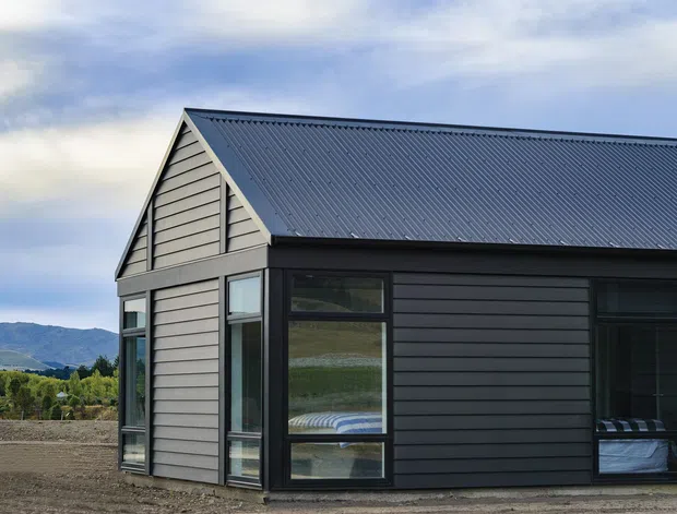 Exterior view of a compact modern gable-roofed dwelling with dark horizontal cladding, a corrugated metal roof, and large black-framed windows. The building sits in a rural or semi-rural landscape with mountains visible in the background.