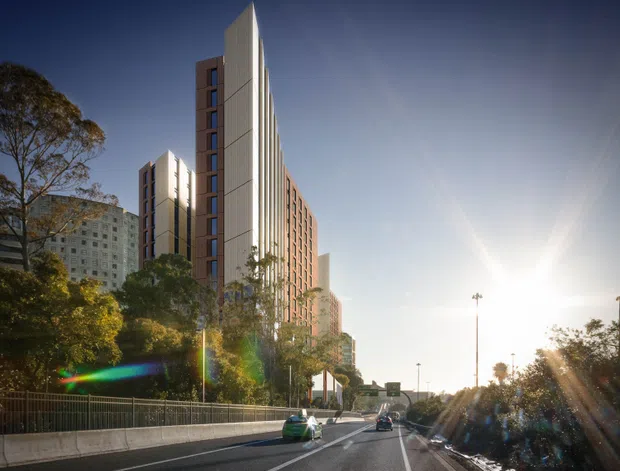 Street-level exterior view of a tall contemporary multi-storey residential or mixed-use building with a sharp, sculptural vertical profile and pale ribbed facade panels, alongside a lower brick-toned wing. The building rises behind landscaped roadside trees and infrastructure, with a roadway and bright low sun in the foreground.
