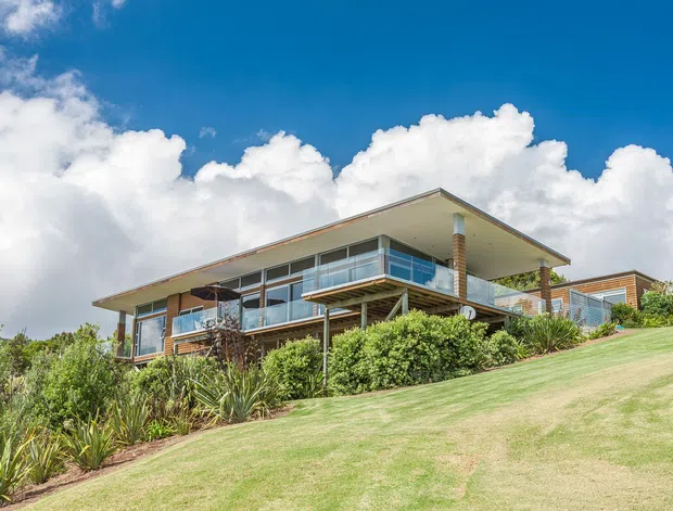 Exterior view of a contemporary multi-level home set on a sloping landscaped site. The building features broad low-pitched roof planes with deep eaves, extensive glazing, timber-toned wall sections, and elevated balconies with clear glass balustrades. Dense garden planting and a manicured lawn surround the house beneath a bright blue sky.