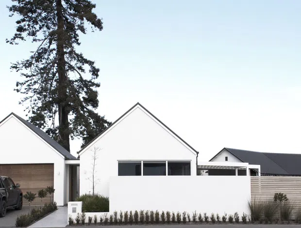 Street-level exterior view of a contemporary residential development with crisp white gabled façades, dark pitched roofs, timber-toned garage doors, and a low privacy wall with landscaped planting. A driveway, garage, and parked vehicle are visible in the foreground, with mature trees behind the homes.