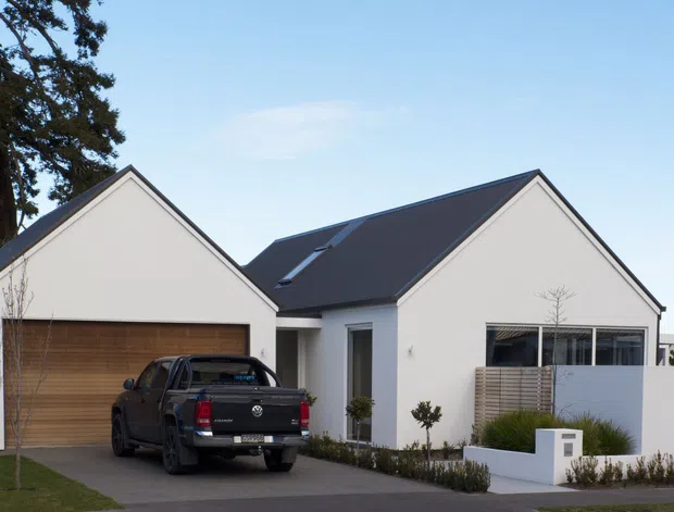 Contemporary single-storey suburban house with two prominent gabled roof forms, pale rendered walls, dark metal roofing, a timber garage door, and a partially screened front courtyard. A pickup truck is parked in the driveway, with low planted landscaping around the entry and boundary walls.