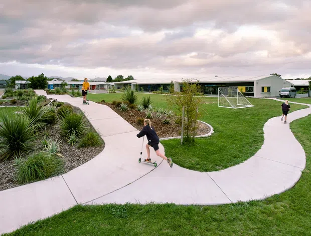 Wide exterior view of a low, modern single-storey community or school-like building set within landscaped grounds. Curving concrete pedestrian paths wind through ornamental grasses and planted garden beds, with children using scooters and a small soccer goal on the lawn. The building has a long horizontal form, pale cladding, extensive glazing, and a low-pitched roof.