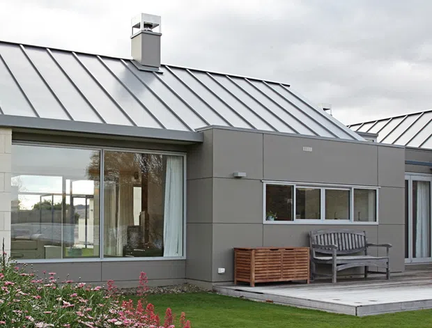 Contemporary single-storey residential house with a low-pitched standing-seam metal roof, pale masonry walls, dark grey cladding to the central extension, broad glazing, and a simple paved outdoor terrace. The composition shows a clean, restrained modern facade with a small landscaped lawn and garden planting.
