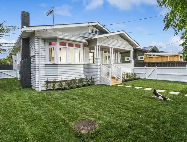 Exterior view of a renovated single-storey weatherboard bungalow with a gabled roof, covered front porch, projecting bay window, dark brick chimney, white picket-style fencing, and a neatly landscaped lawn with stepping stones.