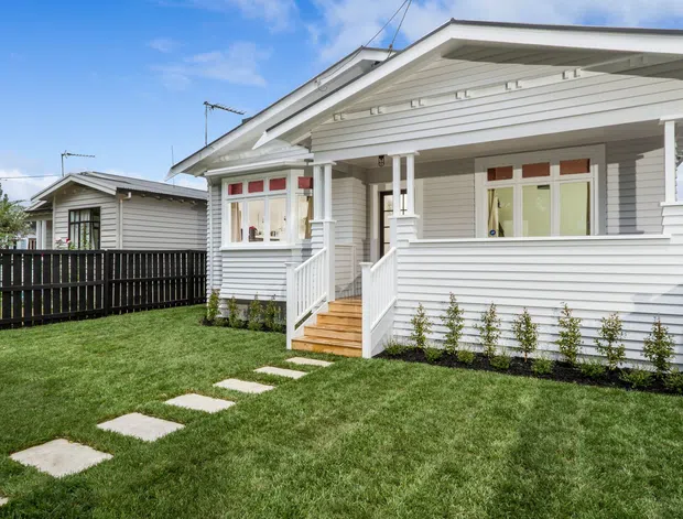 A bright exterior view of a renovated single-storey weatherboard home with a prominent covered front veranda, white timber detailing, steps, sash-style windows, and a neatly landscaped lawn with stepping stones. A second matching dwelling and dark timber fence are visible behind it.