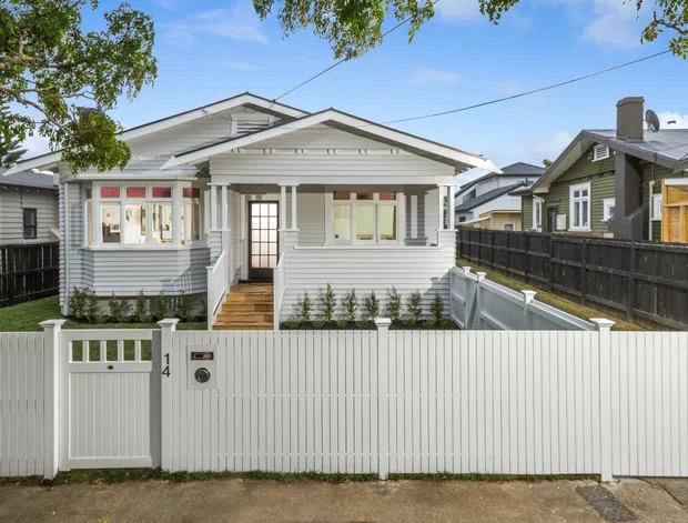 Street-facing view of a compact single-storey weatherboard bungalow with a light painted facade, gabled roof, covered front porch, bay window, white picket-style privacy fencing, and a small landscaped front garden. Neighbouring homes and overhead utility wiring are visible in the suburban setting.