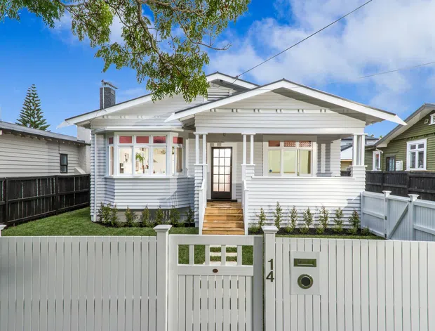 Front exterior view of a single-storey weatherboard bungalow with a prominent gabled verandah, white painted cladding, decorative timber trims, a small landscaped front yard, and a white picket-style boundary fence and gate.