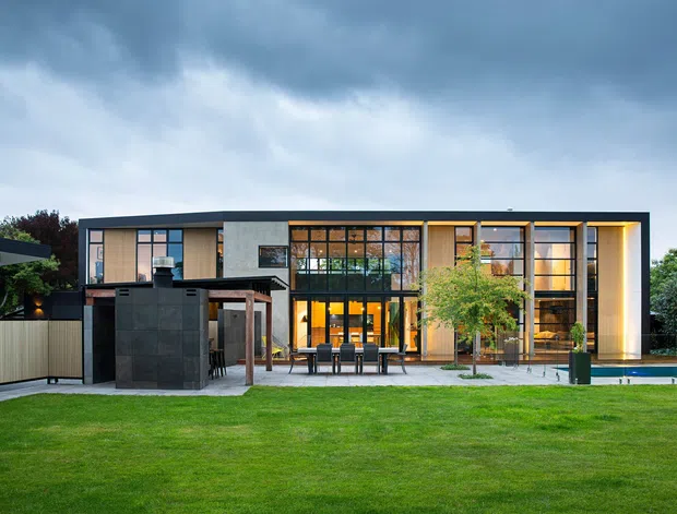 A contemporary two-storey residence viewed from the landscaped rear garden. The home has a strongly horizontal flat-roofed form, extensive floor-to-ceiling glazing with dark metal framing, warm timber-toned interior surfaces, pale masonry or concrete wall panels, and a covered outdoor entertaining area beside a narrow swimming pool. A dark outdoor kitchen or bar structure sits in the foreground, with lawn and planting surrounding the house.