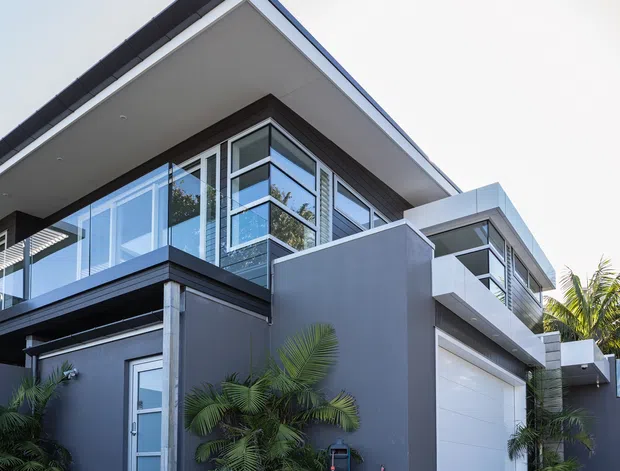 Contemporary multi-level residential house exterior with a sharply articulated flat roof, dark grey rendered walls, extensive corner glazing, white roof soffits and trim, and a large white sectional garage door. Tropical palms and planted hedging frame the facade, while balconies and projecting upper-level volumes create a modern geometric composition.
