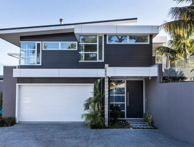 Front exterior view of a contemporary two-storey residence with dark horizontal cladding, white rendered and panelled elements, extensive glazing, a flat low-pitched roof profile, integrated double garage, and a recessed entry. Tropical palms and planting frame the driveway.