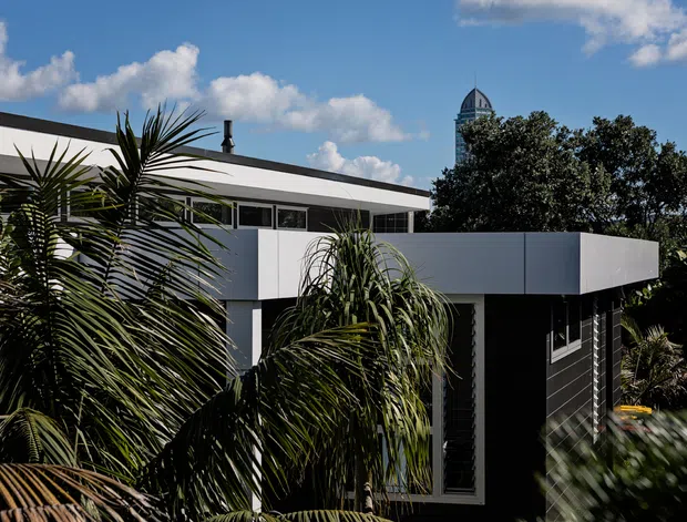 Exterior view of a contemporary flat-roofed residence, with crisp white and dark facade elements, horizontal glazing, and lush tropical planting in the foreground. A distinctive tower is visible in the background under a bright blue sky.