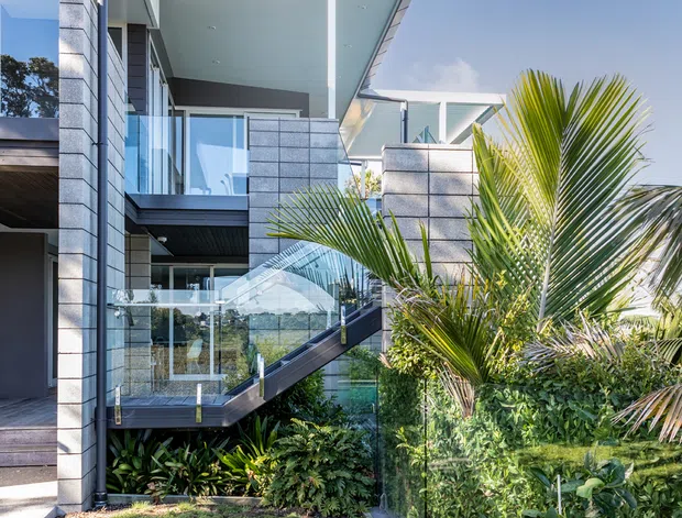 Bright exterior view of a contemporary multi-level residence with flat projecting roof planes, pale masonry cladding, extensive glazing, glass balustrades, and a projecting balcony or stair structure. Tropical planting and a manicured lawn frame the modern house.