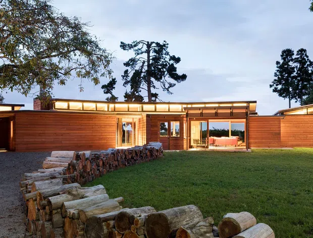 Single-storey contemporary timber-clad residence set within a landscaped suburban or rural site. The low horizontal building has extensive glazing, a shallow roofline with clerestory-like upper windows, a covered carport at the left, and warm interior lighting visible through the central entrance glazing. A rustic stacked-log retaining or boundary feature leads across the lawn in the foreground.