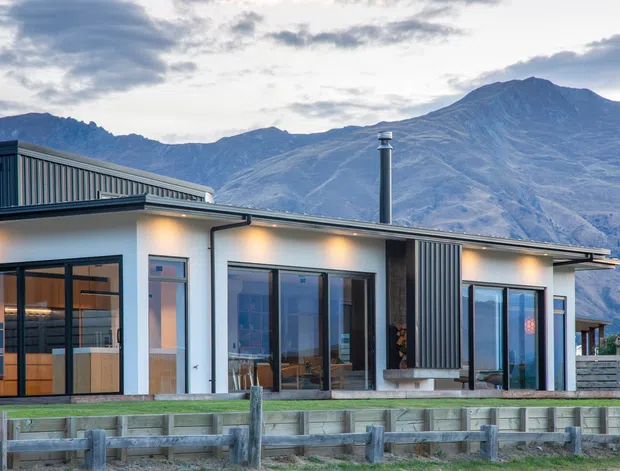 A contemporary single-storey house with a low-pitched roof, white rendered walls, dark vertical metal cladding accents, extensive glazing, and warm exterior lighting. The property is viewed from outside with a timber post-and-rail fence in the foreground and dramatic mountains behind it.