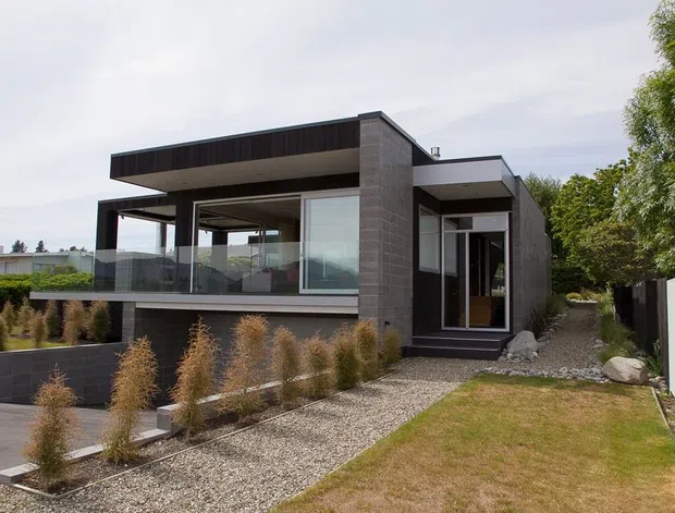 A contemporary two-storey residence with a strongly rectilinear form, dark vertical cladding, concrete-toned wall surfaces and extensive floor-to-ceiling glazing. The upper level includes a cantilevered covered terrace or balcony enclosed by clear glass balustrades, while a gravel path, structured planting and lawn frame the front approach.