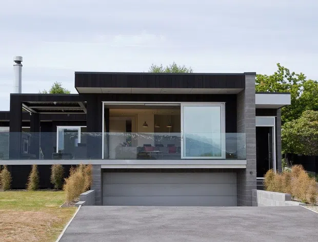 Front exterior view of a contemporary single-storey residence with a dark cladding and masonry facade, broad flat roof forms, large glazed openings, a glass-fronted upper terrace or balcony, and an integrated double garage. The landscaped frontage includes ornamental grasses and a paved driveway.