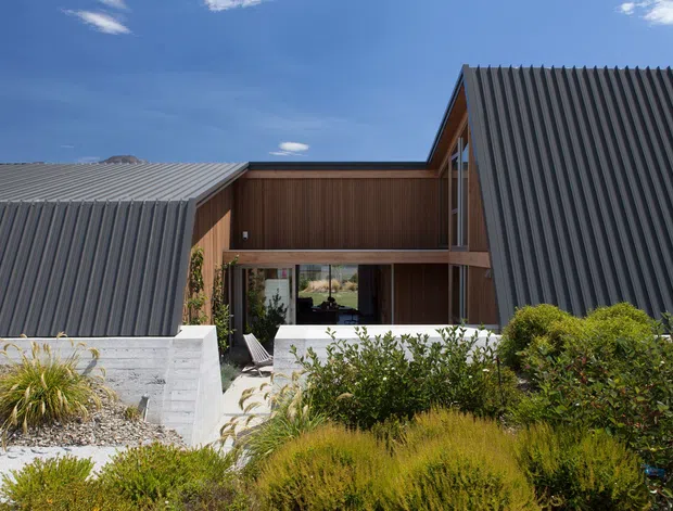 Exterior view of a contemporary, low-rise house composed of sharply pitched dark ribbed metal roof forms around a recessed central glazed entry or courtyard. Warm vertical timber cladding frames the central section, with pale concrete retaining walls and structured native-style landscaping in the foreground. Blue sky and rugged terrain are visible beyond the roofline.