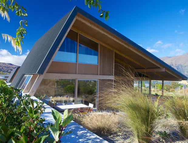 A contemporary A-frame house set in a mountainous landscape, viewed from the landscaped front approach. The home features a steeply pitched gable roof with dark metal roofing, extensive floor-to-ceiling glazing, warm timber soffits and wall cladding, and a partially covered outdoor terrace. Ornamental grasses, shrubs, and a paved walkway frame the foreground, with rugged mountains visible beyond.