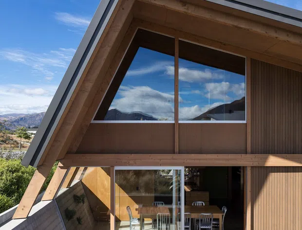 Exterior view of a contemporary two-storey timber-clad house with a steep asymmetrical gable roof, large glazed openings, and a concrete outdoor terrace furnished with a timber dining table and chairs. The building sits in a mountainous landscape, with extensive timber soffits, vertical timber wall cladding, and strong indoor-outdoor visual connection.