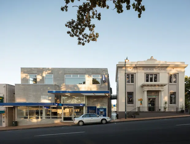 Street-level exterior view of a commercial urban streetscape, showing a modern low-rise building beside a prominent neoclassical bank façade with columns, decorative cornice, pilasters, and the inscription “Bank of New Zealand.” Retail or commercial frontage occupies the ground level, with a broad paved roadway in the foreground and mature tree canopy above.