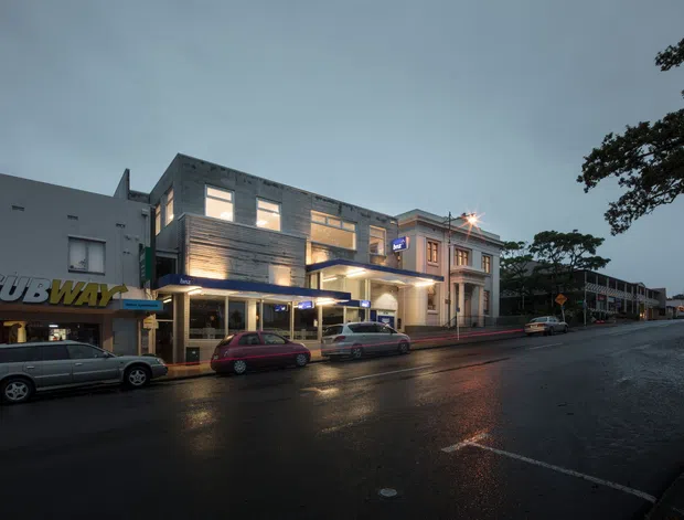 Twilight street-level photograph of a mixed-use urban commercial building on a wet road. The main building has a contemporary multi-storey facade with horizontal timber-like cladding, large illuminated windows, projecting balconies and a sheltered ground-floor frontage. Adjacent retail and older commercial buildings line the street, including a visible Subway storefront. Streetlights and vehicle reflections create a subdued evening atmosphere.
