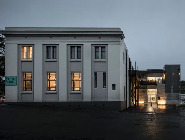 Exterior dusk view of a renovated multi-storey building. The foreground is a formal grey rendered facade with tall vertically proportioned windows, recessed panels, and a projecting classical cornice. A contemporary addition with dark cladding, exposed structural elements, glazing, and illuminated circulation space is visible to the right. Wet paving and subdued evening light create a moody atmosphere.