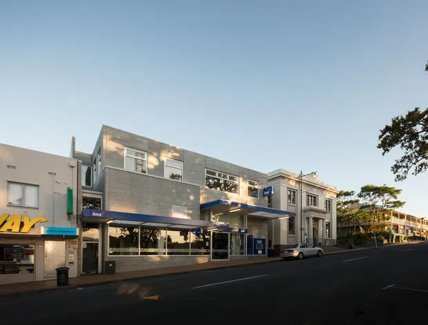 Street-level view of a predominantly modern commercial building within an urban shopping or business streetscape. The main building has a grey brick and concrete facade, extensive ground-floor glazing beneath a projecting canopy, upper-level windows, and a small blue projecting sign. Older decorative commercial architecture is visible on the adjoining building, with additional low-rise shopfronts extending along the street.