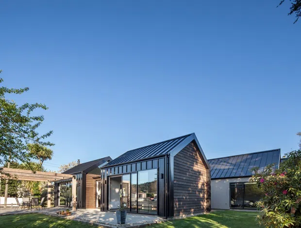 Exterior view of a contemporary single-storey residence composed of several connected gabled volumes. The home features dark standing-seam metal roofs, dark timber weatherboard cladding, extensive black-framed glazing, and a covered timber outdoor entertaining structure. The landscaped foreground includes lawn and planting under a clear blue sky.