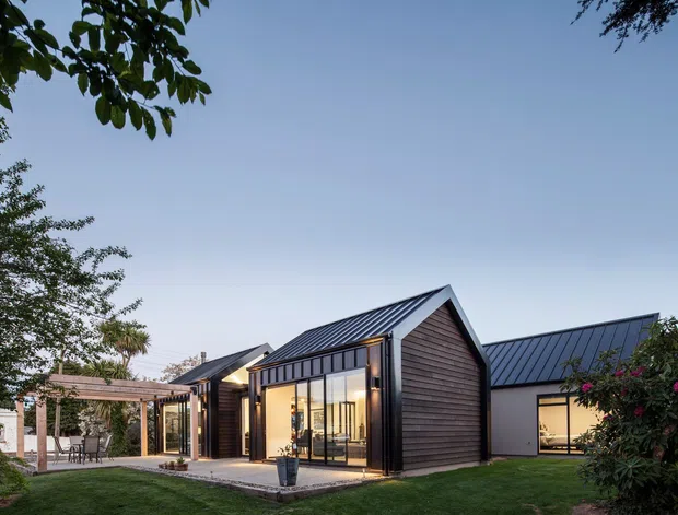 Exterior view of a contemporary single-storey residence composed of connected gabled volumes. The home features dark vertical-seam metal pitched roofs, weathered timber cladding, extensive floor-to-ceiling glazing, and a sheltered timber pergola opening onto a landscaped lawn and paved terrace. Warm interior lighting is visible through the windows at dusk.