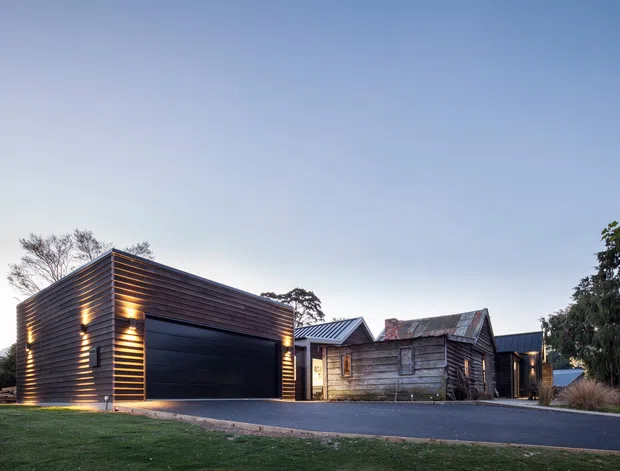 Exterior architectural photograph of a modern dark timber-clad garage or outbuilding beside a weathered timber-clad heritage-style structure. The composition shows horizontal wood cladding, a large black sectional garage door, pitched metal roofs, recessed exterior lighting, and a broad paved driveway, set within a landscaped suburban setting at dusk.