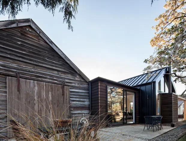 Exterior view of a contemporary dark-clad extension connected to a weathered timber barn or shed. The composition shows a pitched metal roof, large glazed openings, dark vertical cladding, and a paved outdoor seating area, with the original rustic timber structure forming the left side of the image.
